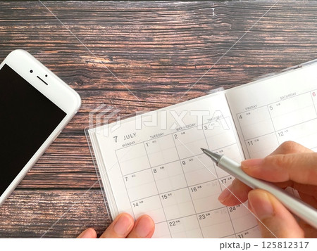 机上で7月のスケジュール帳に記入する手元とスマホ 机上で7月のスケジュール帳に記入する手元とスマホ 125812317