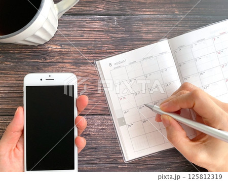 スマホ片手に8月のスケジュール帳に記入する手元 スマホ片手に8月のスケジュール帳に記入する手元 125812319