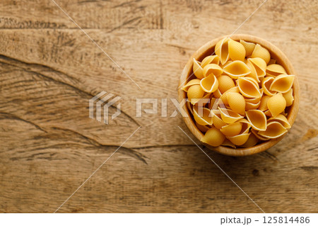Raw uncooked dried Conchiglie shell pasta. Italian seashells pasta in wooden bowl on wooden background. Raw uncooked dried Conchiglie shell pasta. Italian seashells pasta in wooden bowl on wooden background. 125814486