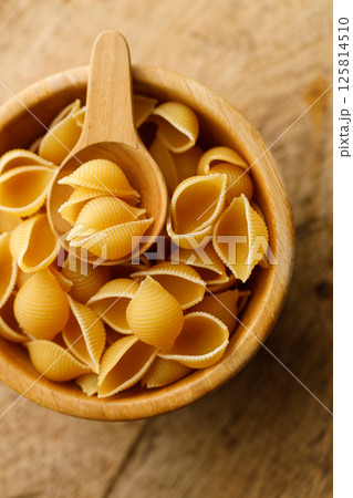 Raw uncooked dried Conchiglie shell pasta. Italian seashells pasta in wooden bowl on wooden background. Raw uncooked dried Conchiglie shell pasta. Italian seashells pasta in wooden bowl on wooden background. 125814510