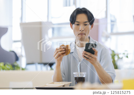 ハンバーガーを食べながらスマートフォンを操作する30代の男性 125815275