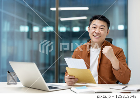 An asian man is happy and celebrating a success, holding documents at his desk. A laptop and pen holder are visible. The background shows a modern office. An asian man is happy and celebrating a success, holding documents at his desk. A laptop and pen holder are visible. The background shows a modern office. 125815648