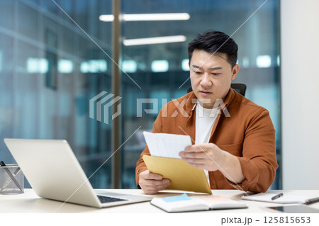 Asian man dressed casually examining documents at a modern office desk, with a laptop, notebook, and stationary, portraying a professional and focused demeanor. 125815653