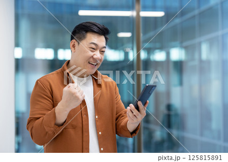 Successful Asian man standing with phone in hands inside office at workplace. Businessman celebrating successful achievement results, received online notification on smartphone about triumphant win. Successful Asian man standing with phone in hands inside office at workplace. Businessman celebrating successful achievement results, received online notification on smartphone about triumphant win. 125815891