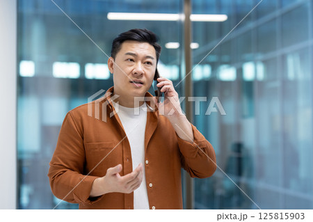 Asian man mature businessman in an office setting, engaged in a conversation using a smartphone. The image captures a professional moment, reflecting communication, business, and technology use Asian man mature businessman in an office setting, engaged in a conversation using a smartphone. The image captures a professional moment, reflecting communication, business, and technology use 125815903