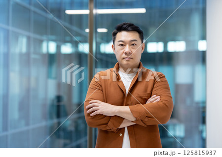 Serious mature man with crossed arms inside office at workplace looking at camera. Successful asian businessman investor, in casual clothes standing near window. Serious mature man with crossed arms inside office at workplace looking at camera. Successful asian businessman investor, in casual clothes standing near window. 125815937