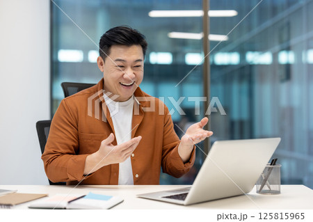 A happy asian man is sitting in an office, smiling and gesturing during a video call on his laptop. Modern office setting. 125815965