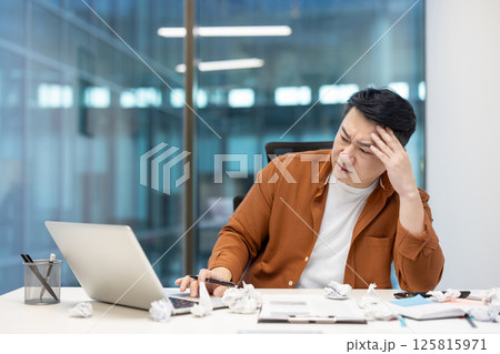 An asian businessman looks stressed while working at his desk, with crumpled paper surrounding him, symbolizing writer's block. 125815971