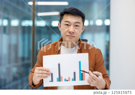 Video call, online conference presentation. Man looks into the camera and shows colleagues a graph with income and financial indicators. Businessman at workplace inside the office. 125815973