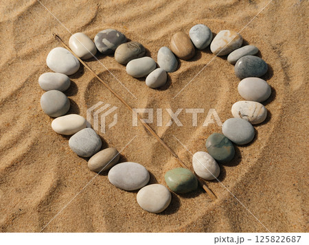 Heart shape made from smooth beach pebbles on sand with a wooden stick crossing it, creating a peaceful natural composition and romantic summer atmosphere perfect for design and decoration themes Heart shape made from smooth beach pebbles on sand with a wooden stick crossing it, creating a peaceful natural composition and romantic summer atmosphere perfect for design and decoration themes 125822687