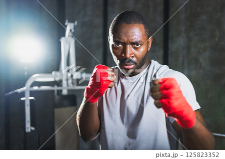 Workout in gym. African man fighter ready for fight punching with boxing wraps protective bandages to camera. Strong man training punches looking concentrated straight preparing for boxing sparring 125823352