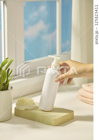 A slender hand tilts a bottle of cosmetics. A hand comb sits on a rectangular stone slab, next to a cactus pot. Light from the window shines in the scene, shadows of objects fall on the white table. A slender hand tilts a bottle of cosmetics. A hand comb sits on a rectangular stone slab, next to a cactus pot. Light from the window shines in the scene, shadows of objects fall on the white table. 125823411