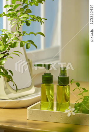 Two transparent green cosmetic bottles in travel sample form, placed on a polished stone tray. A table mirror placed on a tray behind, along with a lush houseplant. A few daisies decorated around. Two transparent green cosmetic bottles in travel sample form, placed on a polished stone tray. A table mirror placed on a tray behind, along with a lush houseplant. A few daisies decorated around. 125823414