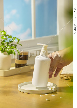 The shampoo bottle was held lightly in the air by a hand. Daisies and flower petals scattered around for decoration. Amber bottles were stacked on top of each other, next to a pot of green leaves. 125823416