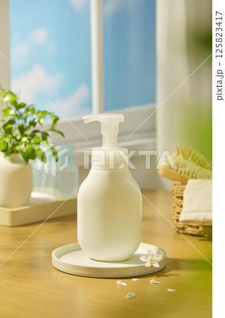 Frontal shot of pump bottle mockup, sitting on a circular stone podium with daisy petals scattered around. Behind are micellar water bottles and indoor plants, placed near the windowsill. 125823417