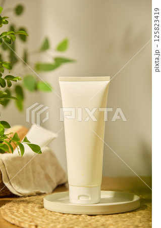 Fingers of hand touch the pump head of a pink cosmetic bottle, filled with liquid and placed on a straw mat, next to a wicker basket containing cosmetic tubes, a wooden comb and coarse cloth. 125823419