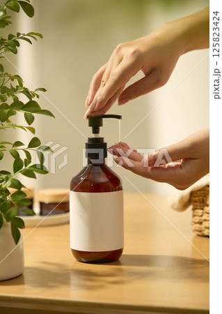 A hand gently presses the pump nozzle of the bottle, the essence flows out from the amber bottle, another hand is catching the essence. Frontal shot of an unlabeled bottle, used for advertising. 125823424