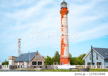 Rustic red-and-white Pakri lighthouse in Estonia stands tall against a bright blue sky, surrounded by small cozy buildings and lush greenery 125823686