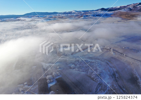 Aerial view of snow-covered landscape with fog rolling over fields and hills in winter season 125824374