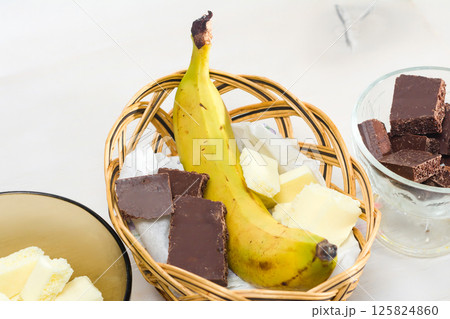 Banana, dark and white chocolate pieces in a wicker basket and glass bowls. 125824860