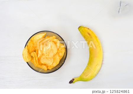Potato chips in a bowl and a banana on a white wooden table. Potato chips in a bowl and a banana on a white wooden table. 125824861