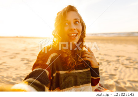 Young woman taking a cheerful selfie on a sandy beach during sunset. Lifestyle, travel, active life 125825142