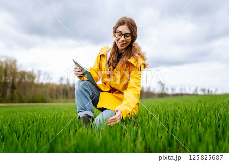 Young farmer woman in a green field, intently using a tablet. She wears a yellow jacket and glasses 125825687