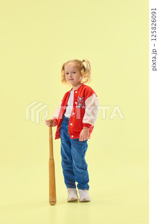 Smiling girl in red varsity jacket holds wooden baseball bat, ready to play against yellow background. Smiling girl in red varsity jacket holds wooden baseball bat, ready to play against yellow background. 125826091
