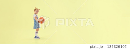 Cute little girl in denim overalls holding basketball, standing in studio with calm expression, ready for game on yellow background. Cute little girl in denim overalls holding basketball, standing in studio with calm expression, ready for game on yellow background. 125826105