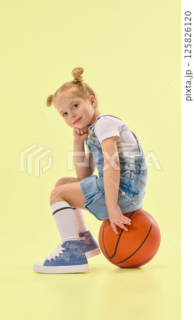 Stylish child sits on basketball, resting chin on hand with thoughtful pose, wearing denim outfit and striped socks in yellow studio background 125826120
