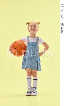 Confident girl smiling, holding basketball on hip, wearing denim overall dress and standing proud, showing positive expression on yellow background. 125826121