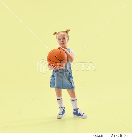 Young girl holds basketball, wearing denim dress and white socks, looking curious and calm, posing against yellow backdrop 125826122