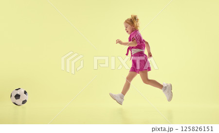 Joyful child girl in pink shorts and shirts running towards soccer ball, playing against yellow background. Joyful child girl in pink shorts and shirts running towards soccer ball, playing against yellow background. 125826151