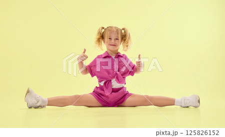 Happy girl child in pink summer clothes and ponytail hairstyle sitting on twine on floor, giving thumbs up and smiling 125826152