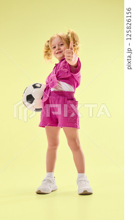 Low angle view of happy child girl in pink shorts and shirt, standing with soccer ball and giving thumbs up against yellow background. Low angle view of happy child girl in pink shorts and shirt, standing with soccer ball and giving thumbs up against yellow background. 125826156