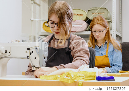 Two female seamstresses in the workshop at the factory are working at tables on sewing machines.  125827295