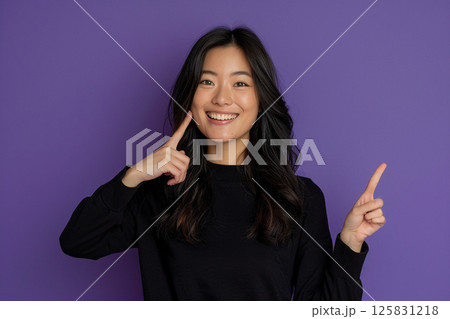 happy young woman pointing up, isolated on a purple background, in side view. Mockup for text and design. Asian girl wearing a white t-shirt, smiling and posing in a studio. 125831218