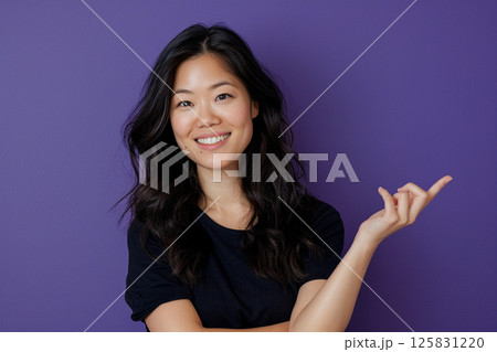 happy young woman pointing up, isolated on a purple background, in side view. Mockup for text and design. Asian girl wearing a white t-shirt, smiling and posing in a studio. 125831220