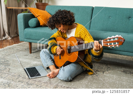 African American woman learning guitar watching tutorial on laptop at home. Black girl playing guitar singing song learning online music lessons. Artistic woman playing acoustic guitar learning chords 125831857