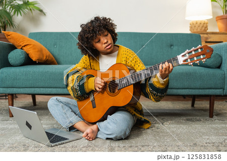 African American woman learning guitar watching tutorial on laptop at home. Black girl playing guitar singing song learning online music lessons. Artistic woman playing acoustic guitar learning chords African American woman learning guitar watching tutorial on laptop at home. Black girl playing guitar singing song learning online music lessons. Artistic woman playing acoustic guitar learning chords 125831858