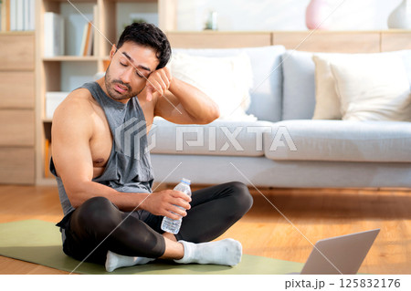 Tired young asian man sitting on a yoga mat after a home workout wiping sweat in living room. 125832176