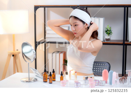 Young asian woman wearing headband and tying hair while preparing skincare looking mirror in room. 125832235