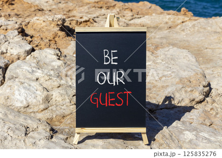 Be our guest symbol. Concept words Be our guest on beautiful blackboard. Beautiful brown stone beach and blue sea sky background. Business and be our guest concept. Copy space. 125835276