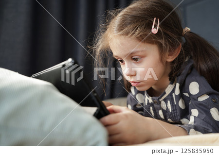 Little girl staring at tablet with intense focus and serious expression while lying on bed Little girl staring at tablet with intense focus and serious expression while lying on bed 125835950