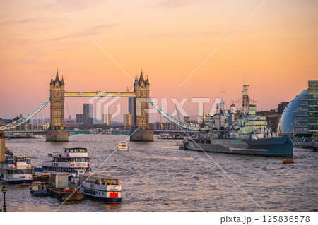Tower Bridge stands majestically over the Thames River as HMS Belfast is docked nearby. The evening sky adds a warm glow to the iconic London skyline, showcasing the city's historical charm. 125836578