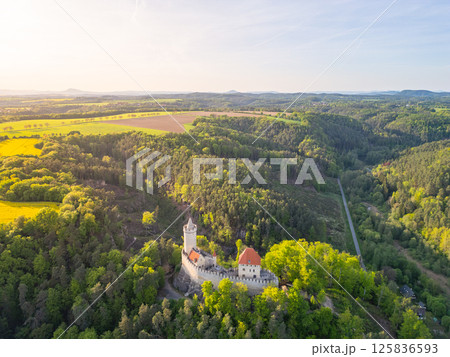 Kokorin Castle stands majestically in a lush green landscape during a sunny evening. The aerial perspective showcases the castle's architectural beauty amid forest and fields. Kokorin Castle stands majestically in a lush green landscape during a sunny evening. The aerial perspective showcases the castle's architectural beauty amid forest and fields. 125836593