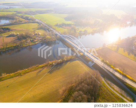Bohumil Hrabal's Bridge gracefully stretches over the Labe River in Litol, Lysa nad Labem, Czechia, surrounded by lush greenery and an expansive landscape during golden hour. 125836624