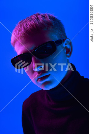 Portrait of stylish young guy in sunglasses and black turtleneck posing against blue background in neon light. Close-up 125836804