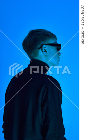Side profile of stylish young man in turtleneck and sunglasses, posing under deep neon blue background ,expresses futuristic elegance. Side profile of stylish young man in turtleneck and sunglasses, posing under deep neon blue background ,expresses futuristic elegance. 125836807
