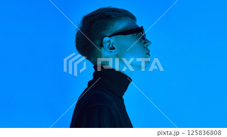 Side profile of stylish young man in turtleneck and sunglasses, posing under deep neon blue background ,expresses futuristic elegance. Side profile of stylish young man in turtleneck and sunglasses, posing under deep neon blue background ,expresses futuristic elegance. 125836808